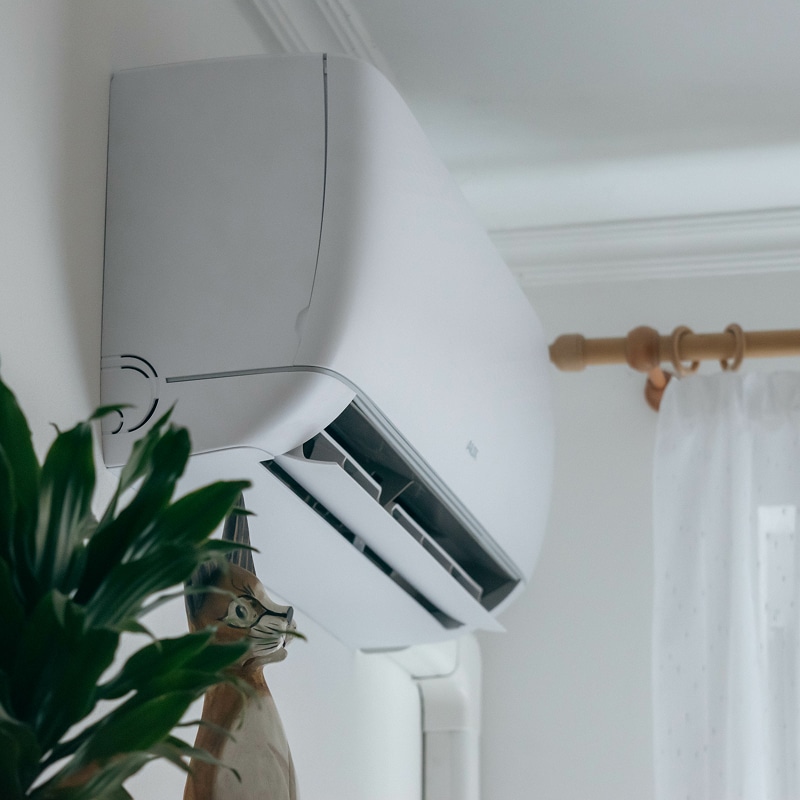 Climatiseur mural blanc AUX avec volets ouverts, installé près d'une plante verte et de rideaux blancs.