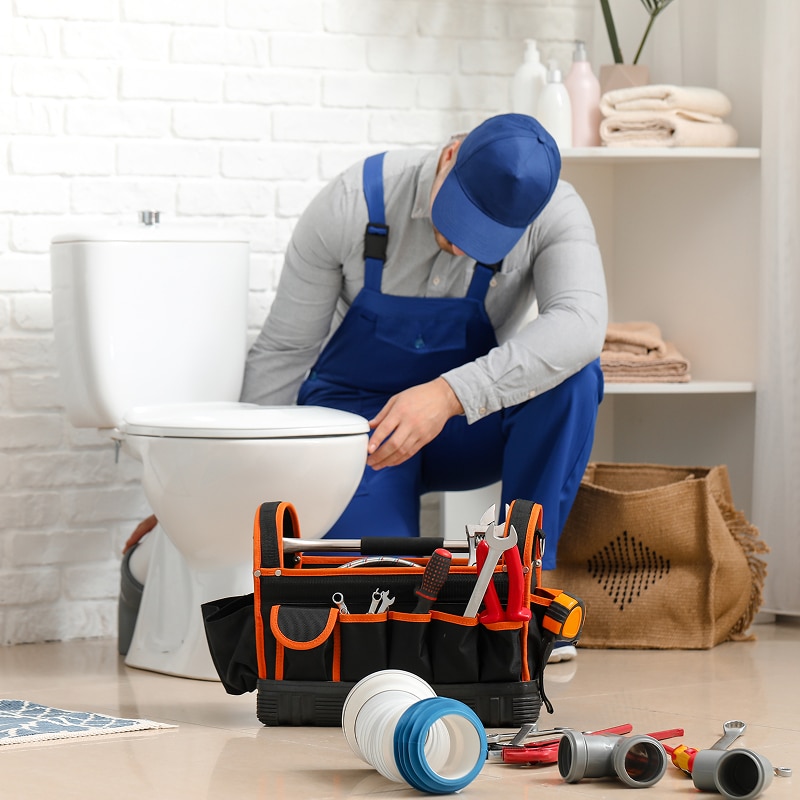 Plombier en salopette bleue travaillant sur une toilette. Boîte à outils pleine d'outils de plomberie au premier plan.